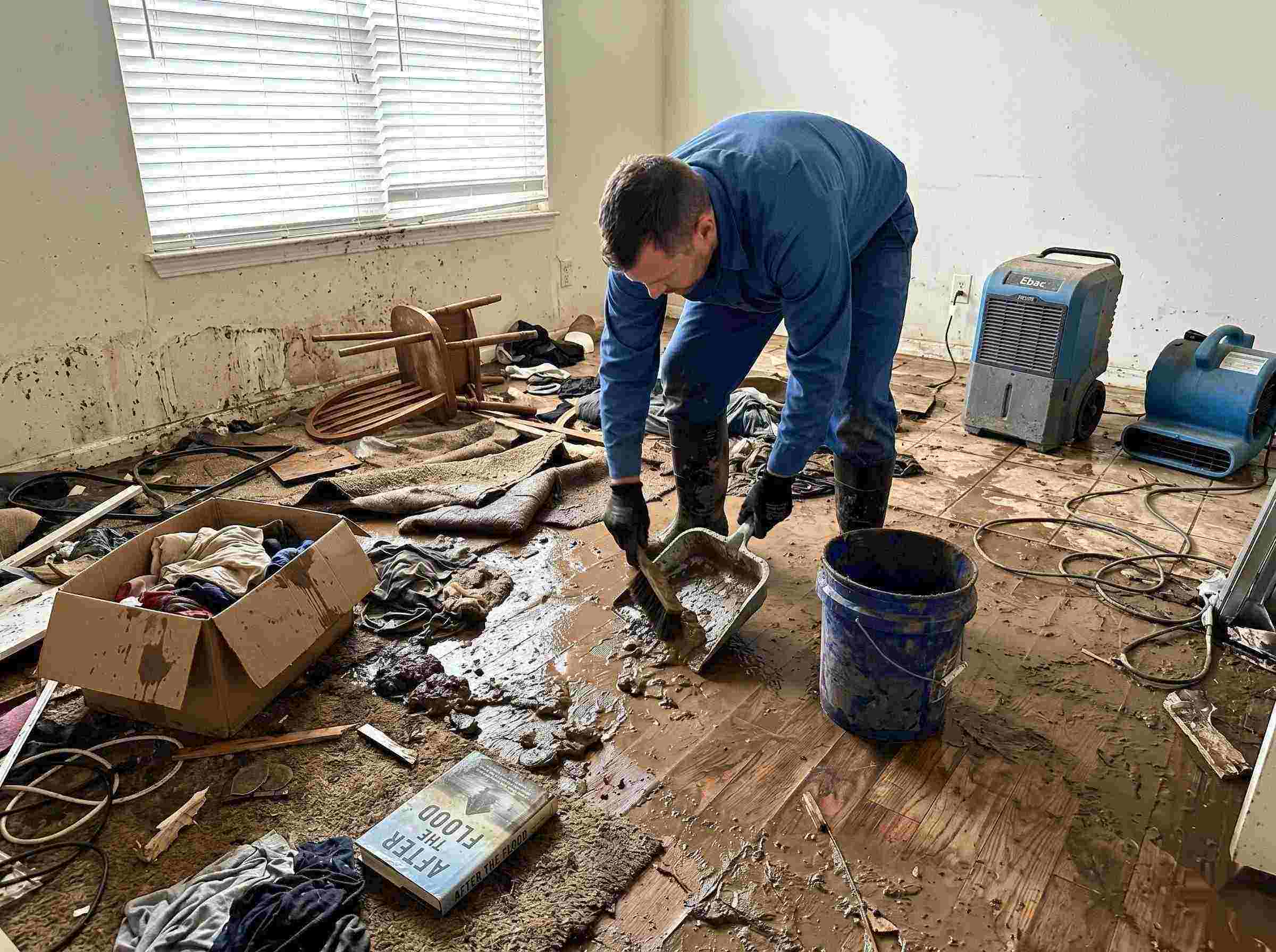 Professional realistic photo of a technician performing mud and debris removal after a home flood in Miramar, showing soaked furniture, wet flooring, and scattered debris during post-flood cleanup.